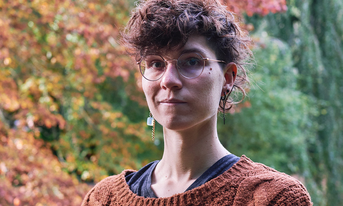 An androgynous white person with wavy dark brown hair, round glasses and ear jewellery.