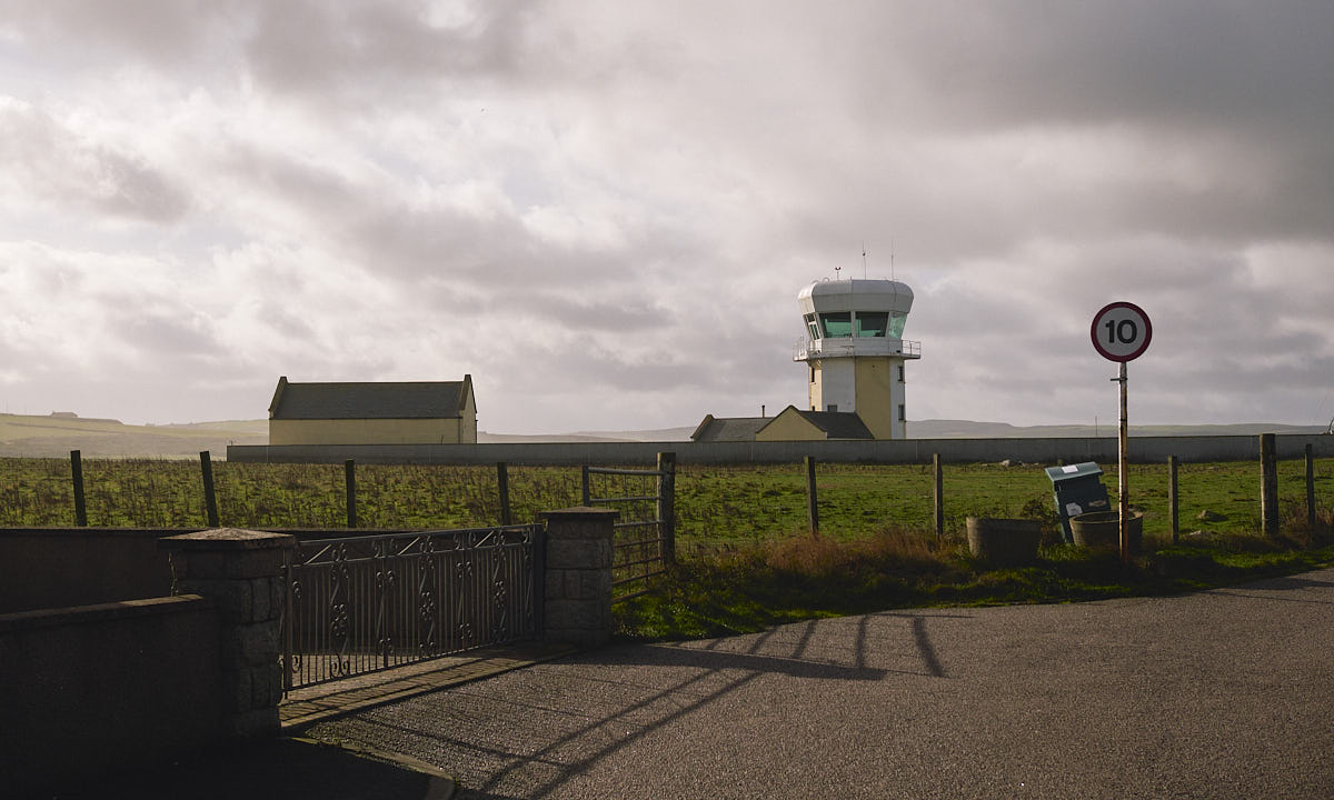 A countryside road with a bin next to a speed sign. In the back, a couple of lone houses and a modern lighthouse stand in a large field