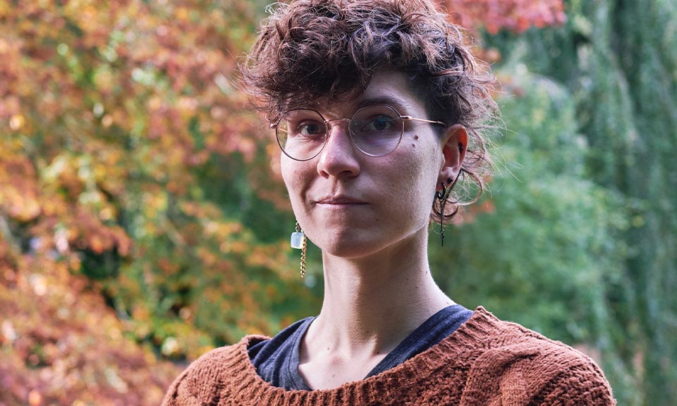 An androgynous white person with wavy dark brown hair, round glasses and ear jewellery.